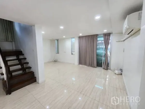 Home Office for rent in Khlong Toei, Bangkok - Bright open-plan living area with glossy tile floors, recessed lights, air conditioner, and a wooden staircase beside sliding doors with curtains.