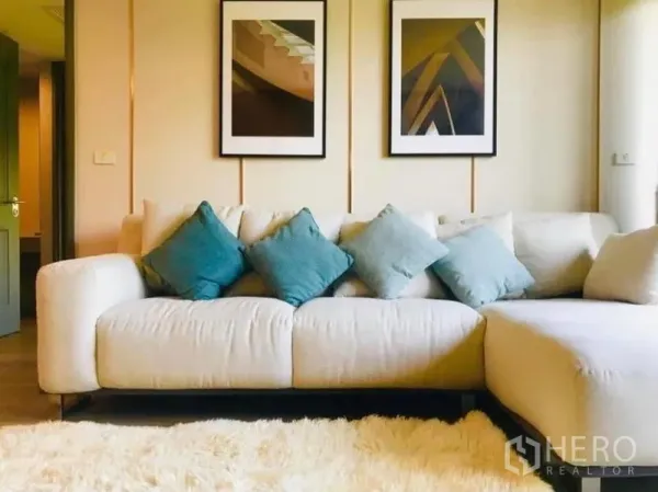 公寓 出租在Pathum Wan, Bangkok - 舒适客厅，米色转角沙发、蓝绿色抱枕、长毛地毯与装饰画。