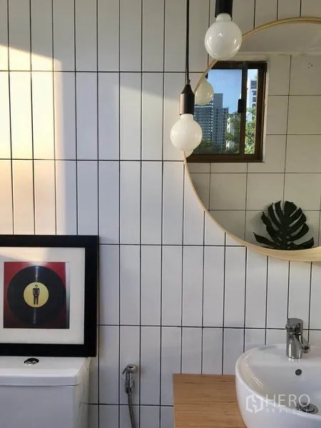 Apartment for rent in Khlong Toei, Bangkok - Bathroom detail showing vertical subway tiles, round mirror, small window, and ceramic sink.