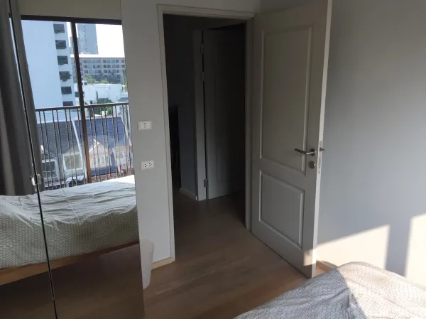 คอนโด ให้เช่าในBangkok - ห้องนอนพื้นไม้ สีอ่อน ประตูเปิดสู่โถงทางเดิน และหน้าต่างใหญ่ติดระเบียง