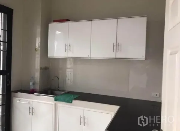 Townhouse for rent in Wang Thonglang, Bangkok - Compact kitchen with white cabinets, black countertop, and sink by a windowed door.