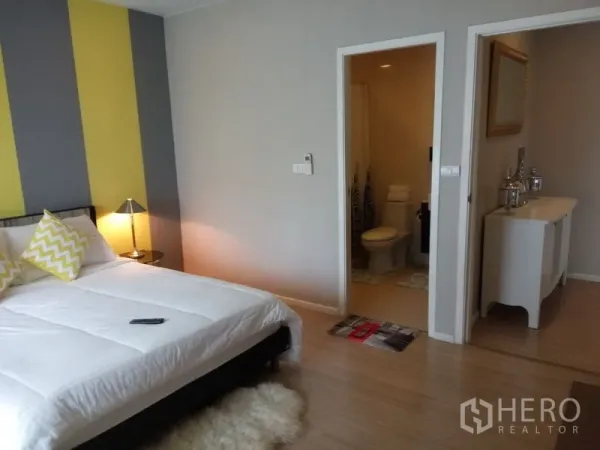 Condominium for rent or sale in Pathum Wan, Bangkok - Bedroom with yellow and gray accent wall, bedside lamps, and en-suite bathroom entry.