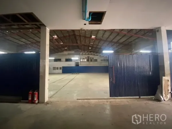 Warehouse for rent or sale in Suan Luang, Bangkok - Entrance view of warehouse showing large blue metal doors, concrete floor and fire extinguishers leading into a spacious hall.
