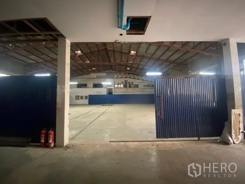 Warehouse for rent or sale in Suan Luang, Bangkok - Entrance view of warehouse showing large blue metal doors, concrete floor and fire extinguishers leading into a spacious hall.