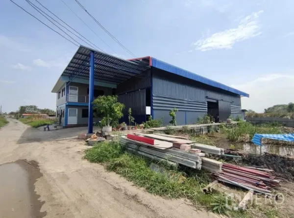 Warehouse for sale in Nakhon Pathom - Side view of the corrugated steel warehouse and office showing roadside access and open yard with stored materials.