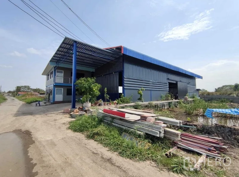 Warehouse for sale in Nakhon Pathom - Side view of the corrugated steel warehouse and office showing roadside access and open yard with stored materials.