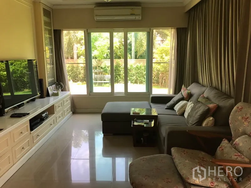 Detached House for sale in Pak Kret, Nonthaburi - Spacious lounge with L-shaped sofa, floor-to-ceiling glass and garden outlook.