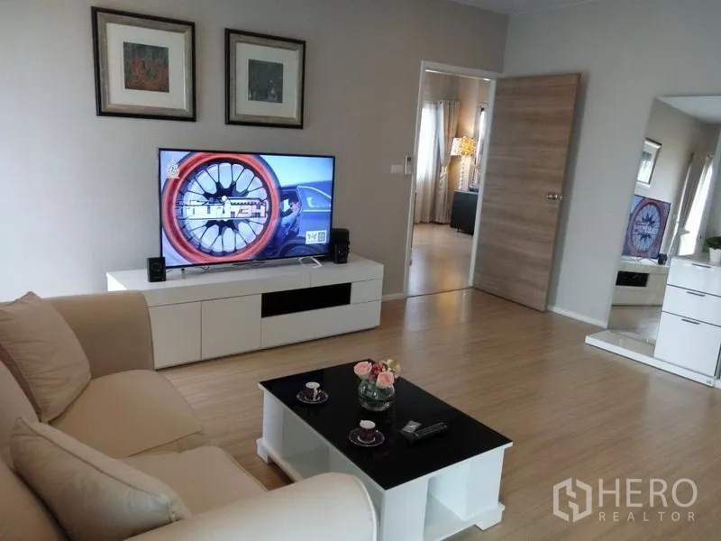 Condominium for rent or sale in Pathum Wan, Bangkok - Cozy family lounge with beige sofa, TV console, wood flooring, and doorway leading to a bedroom.