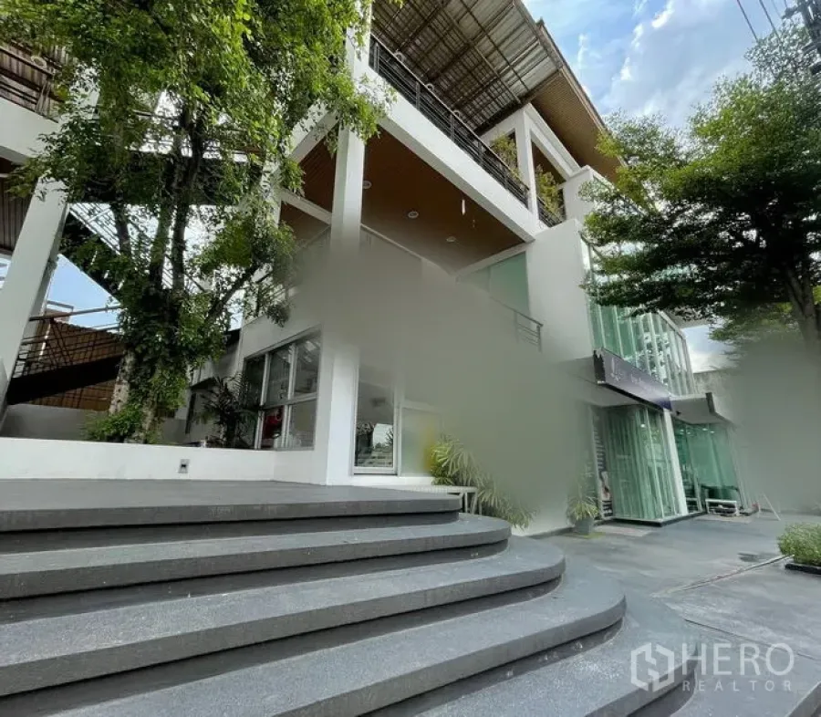 零售空间 出租在Bueng Kum, Bangkok - 现代多层商业建筑，玻璃立面与宽台阶，被大树环绕。