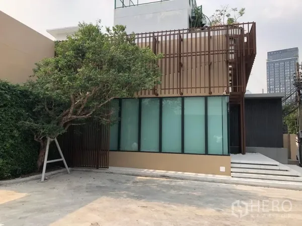 零售空间 出租在Watthana, Bangkok - 临街商铺一角，绿树遮荫的门面、台阶入口与开阔的混凝土停车场。