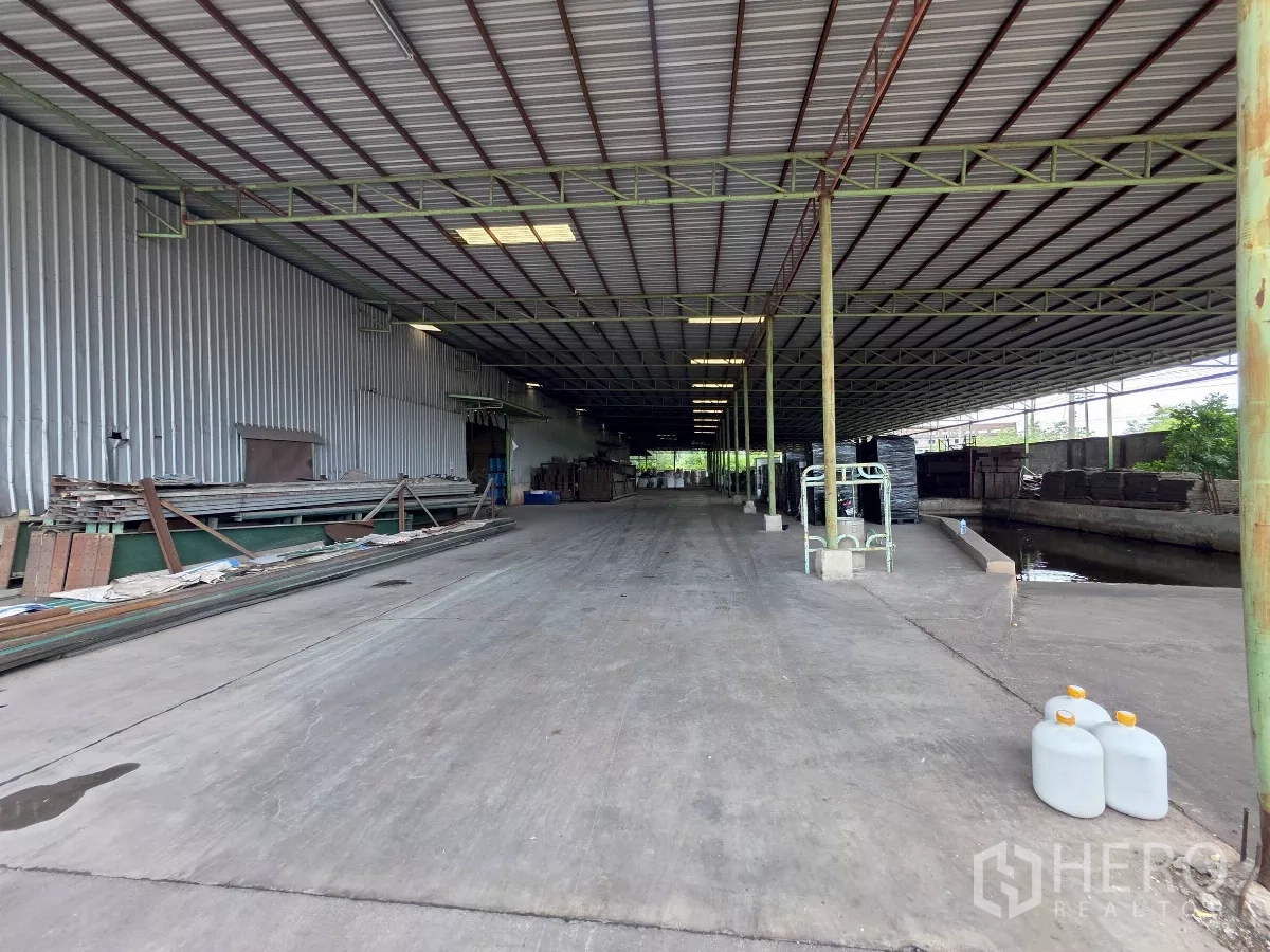 Factory for rent in Mueang Samut Sakhon, Samut Sakhon - Long covered loading bay beside a metal‑clad warehouse with wide concrete driveway and side drainage canal.