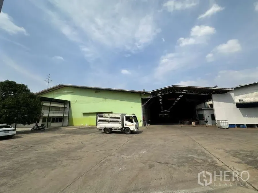 คลังสินค้า ให้เช่าในPak Kret, Nonthaburi - ลานคอนกว้าง พร้อมอาคารโกดังสีเขียวและสีขาว หลังคาทางโหลด และรถบรรทุก