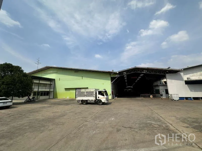 คลังสินค้า ให้เช่าในPak Kret, Nonthaburi - ลานคอนกว้าง พร้อมอาคารโกดังสีเขียวและสีขาว หลังคาทางโหลด และรถบรรทุก