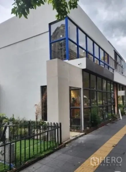 零售空间 出租在Khlong Toei, Bangkok - 两层转角商铺，蓝色框架大玻璃窗与人行道绿化，位于Ekkamai区域。