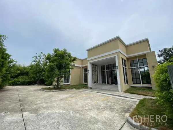 带办公室的家 出租在Bang Na, Bangkok - 现代单层商住两用楼外观，落地大窗与带顶入口，前方宽阔混凝土停车区，周围绿树环绕。