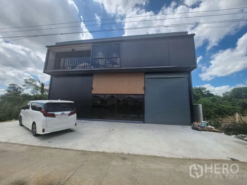 คลังสินค้า ให้เช่าหรือขายในLam Luk Ka, Pathum Thani - ด้านหน้าโกดังสองชั้นโทนสีดำสมัยใหม่ มีระเบียง กระจกบานใหญ่ และประตูม้วน พร้อมลานคอนกรีตจอดรถ