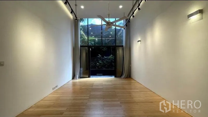 Condominium for rent in Watthana, Bangkok - Long living area with floor-to-ceiling windows, ceiling fan, and curtains opening to a leafy balcony-patio.