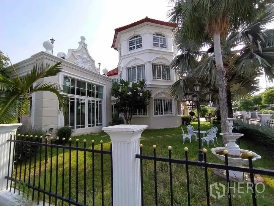Detached House for sale in Phra Samut Chedi, Samut Prakan - Front elevation of an elegant European‑style house with turret, lawn, palms and a garden seating set.