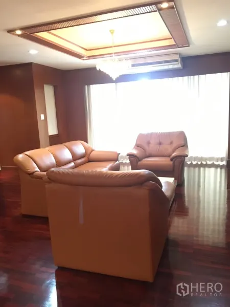Condominium for rent in Watthana, Bangkok - Bright lounge with tan leather sofas beneath a coffered ceiling and chandelier, large window with sheer curtains, and wood floors.