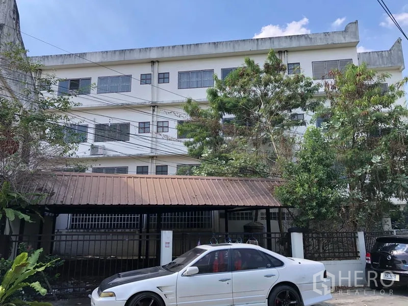 Shophouse for sale in Lam Luk Ka, Pathum Thani - Rear exterior with gated parking area, trees, and a multi-storey building.