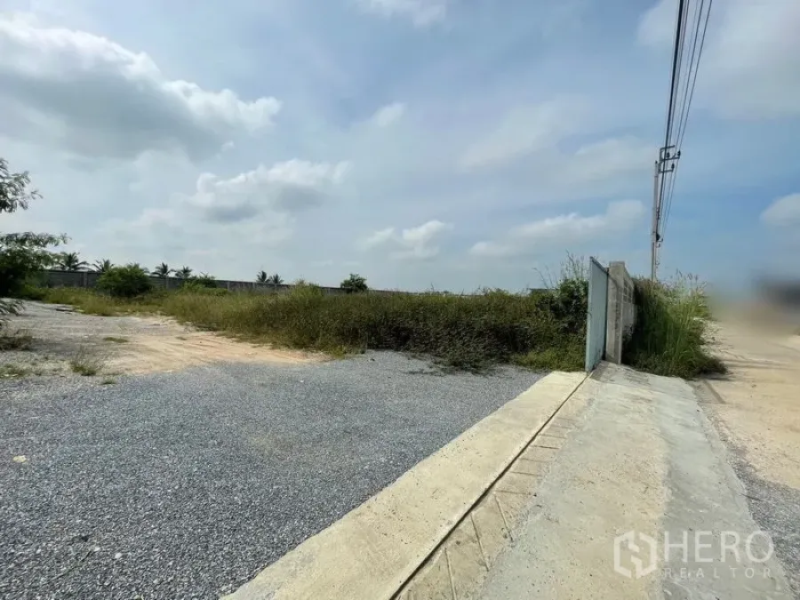 Land for sale in Sai Noi, Nonthaburi - Corner of a vacant plot with gravel entry and a boundary wall along a concrete road in Sai Noi.