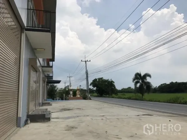 工厂 出租在Pathum Thani - 工厂排屋外观，宽阔混凝土前场，临主干道并望向田野。