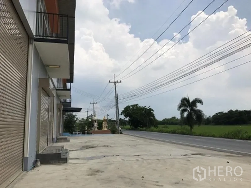 工厂 出租在Pathum Thani - 工厂排屋外观，宽阔混凝土前场，临主干道并望向田野。