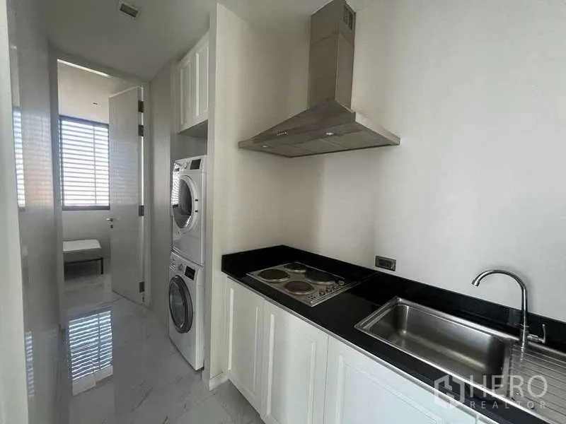 Condominium for rent or sale in Bangkok - Compact utility kitchen with black countertop, cooktop, hood and stacked washer-dryer.