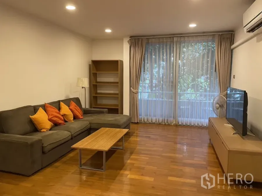 Condominium for rent in Pathum Wan, Bangkok - Bright living room with L-shaped sofa, wood floors, TV console and full-height windows opening to green views.
