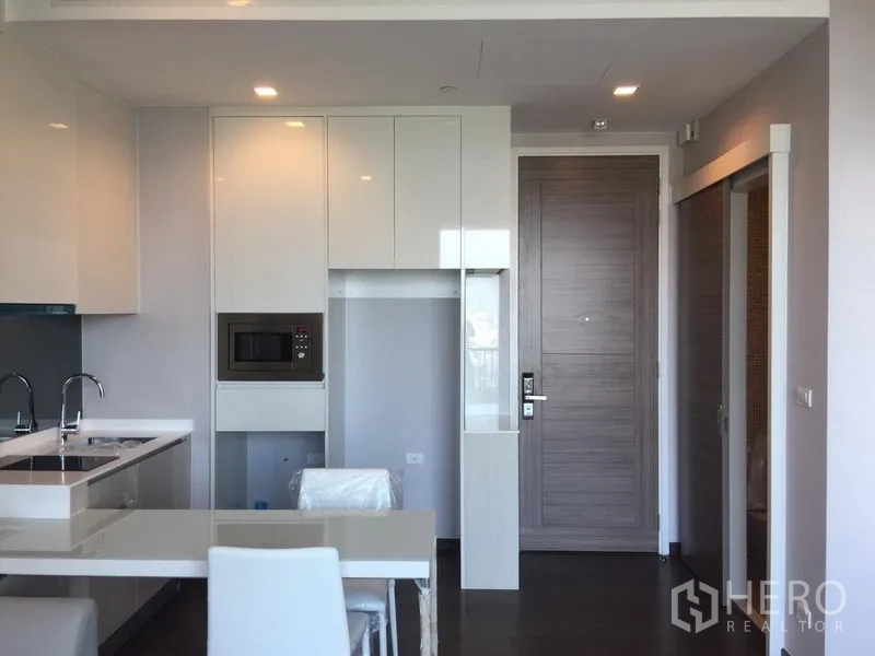 公寓 出租在Watthana, Bangkok - 曼谷公寓的开放式现代厨房，白色亮面橱柜、内置微波炉、餐桌与入户门。