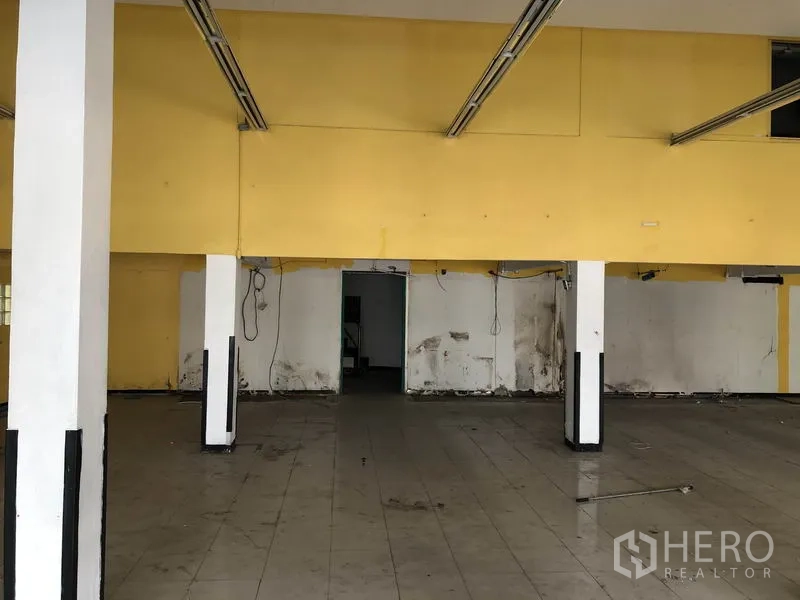 Warehouse for rent in Suan Luang, Bangkok - Wide warehouse hall with ceiling lighting tracks, support pillars, and a central doorway leading to service areas.