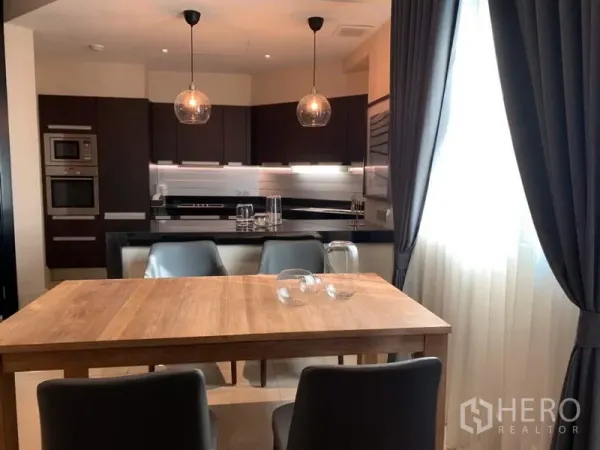 Condominium for rent in Bang Rak, Bangkok - Dining area with a wooden table for four next to a bright window, facing a U-shaped kitchen with pendant lights and built-in appliances.