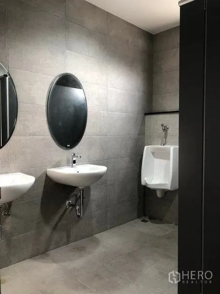 Factory for rent in Samut Prakan - Clean restroom with gray wall tiles, twin wall‑mounted sinks, oval mirrors and a urinal.