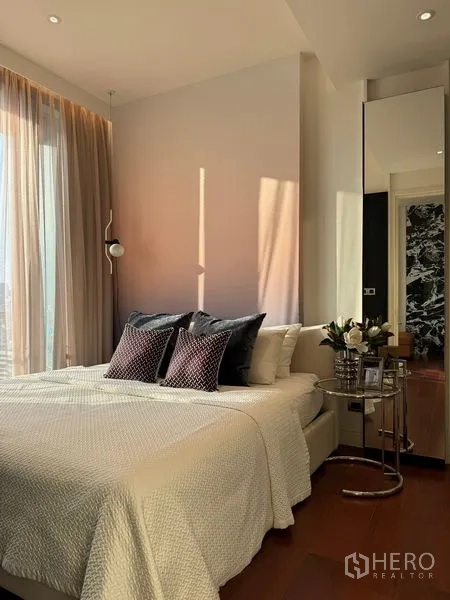 Condominium for sale in Watthana, Bangkok - Bright bedroom with large window, white bedding, accent pillows and a chrome side table with flowers.