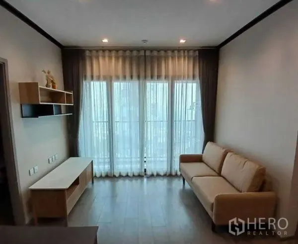 Condominium for rent or sale - Bright living room with floor-to-ceiling windows, sheer curtains, beige sofa, TV console and wood floors.