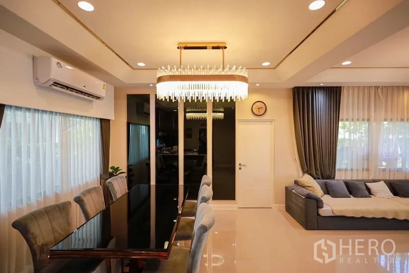 Detached House for rent or sale in Lam Luk Ka, Pathum Thani - Dining room with long table, chandelier and corner sofa by the windows.