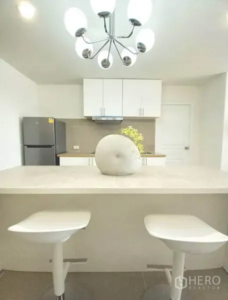 公寓 出租在Khlong Toei, Bangkok - 白色厨房配岛台与两把吧椅，不锈钢冰箱与时尚吊灯。