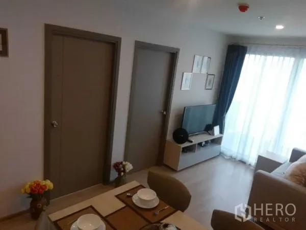 คอนโด ให้เช่าในBang Na, Bangkok - พื้นที่นั่งเล่น–ทานอาหารสว่างโปร่ง มีชั้นวางทีวี ประตูห้องนอนสองบาน และผ้าม่านสูงจรดเพดานที่ระเบียง.