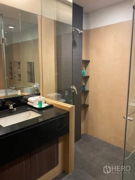 Apartment for rent in Khlong Toei, Bangkok - Vanity with black countertop sink beside a separate glass shower and built-in shelves.