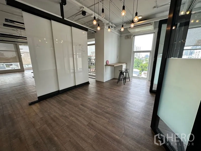 办公空间 出租在Watthana, Bangkok - 明亮的开放式办公角落，木纹地板、玻璃隔断、吊灯、吧台和落地窗城市景观。