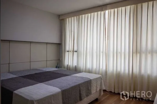 Condominium for rent in Pathum Wan, Bangkok - Bedroom with large curtained window, padded headboard and patchwork duvet on wooden floor