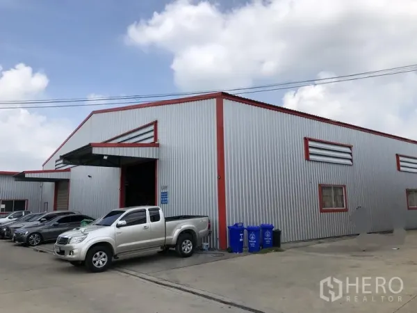 Warehouse for rent in Nonthaburi - Side view of the warehouse facade with large loading door and recycling bins by the driveway.