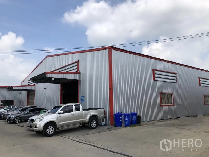 Warehouse for rent in Nonthaburi - Side view of the warehouse facade with large loading door and recycling bins by the driveway.