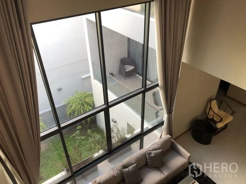 Detached House for rent or sale in Huai Khwang, Bangkok - Double-height living area with floor-to-ceiling windows overlooking an internal courtyard and balcony with greenery.