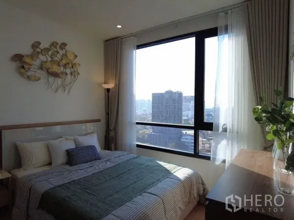 Condominium for sale in Watthana, Bangkok - Sunlit bedroom with wall art, double bed, full-height windows, and urban views.