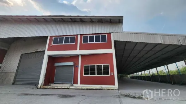 Warehouse for rent in Nonthaburi - Two-storey red office attached to a steel-roof warehouse with a large roller shutter and a wide covered loading bay.