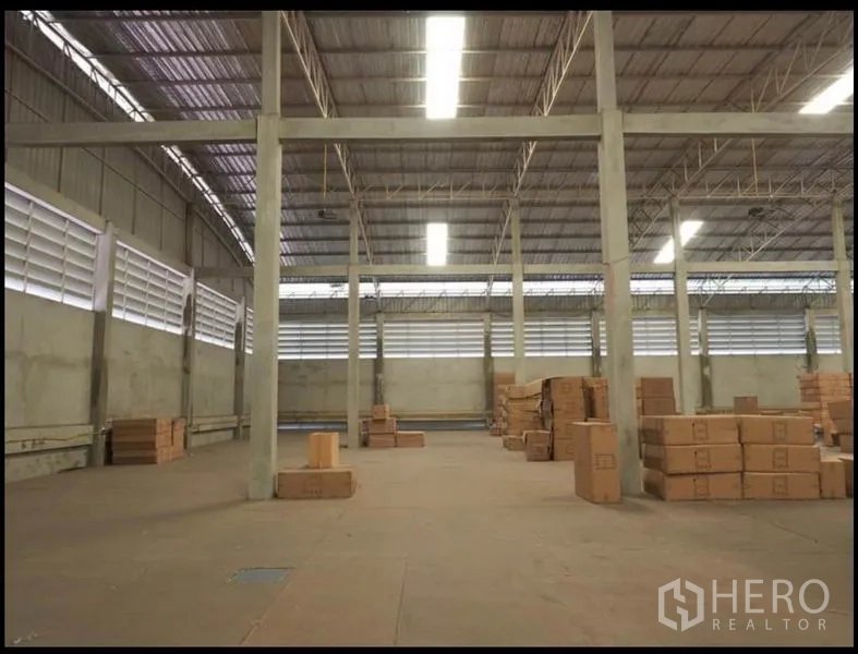 Warehouse for rent in Nakhon Pathom - High-ceiling warehouse hall with louvered side windows, bright strip lighting, and stacked cartons.