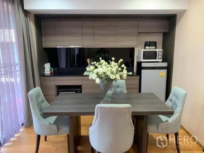 Condominium for rent or sale in Pathum Wan, Bangkok - Modern dining area with four upholstered chairs beside a built-in wood kitchen with fridge and microwave.
