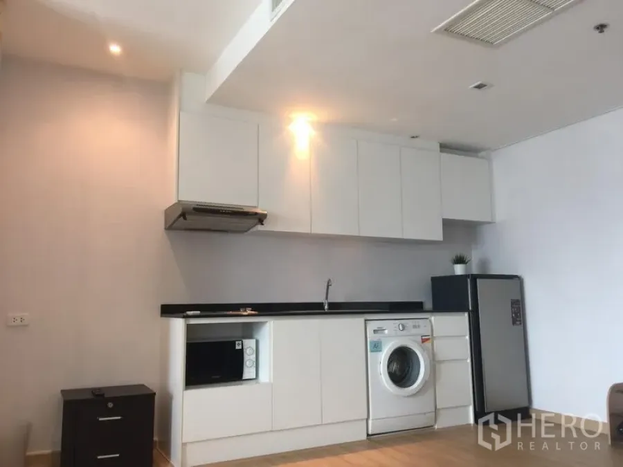 Condominium for rent in Khlong Toei, Bangkok - Full white kitchen wall with cabinets, sink, cooktop, washing machine and refrigerator on wood flooring.