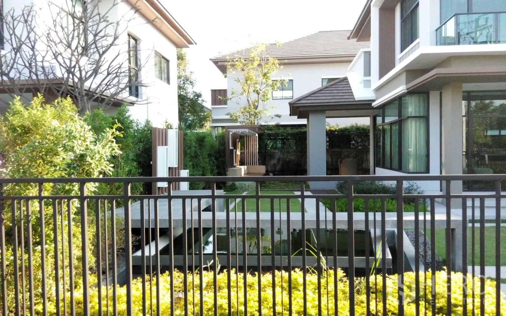 Detached House for rent or sale in Prawet, Bangkok - Landscaped garden with pond and bridge behind a black fence, flanked by neighboring houses.